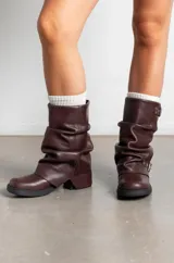 Botas de caña media confeccionadas en cuero color marrón oscuro, con diseño arrugado y dos correas con hebillas metálicas ajustables. Presentan suela plana de goma negra con un pequeño taco cuadrado.