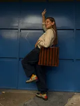 Cartera tipo tote de cuero con diseño de franjas verticales multicolores en tonos tierra, rojo y azul. Cuenta con correas trenzadas de cuero y cierre superior.