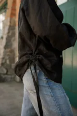 Camisa de lino color negro con silueta relajada, cuello camisero y cierre frontal con botones de nácar. Presenta mangas largas con puños y detalles de tiras laterales.