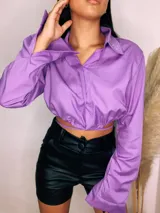 Camisa cropped negra con cuello clásico, mangas largas con puños abotonados y cierre frontal con botones. Presenta detalle de tachas plateadas en el cuello.