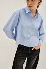 Camisa de corte recto y largo cropped, confeccionada en algodón con estampado de rayas verticales finas en color azul y blanco. Presenta cuello clásico, cierre frontal con botones y mangas largas con puños abotonados. La prenda está decorada con pequeñas aplicaciones de strass distribuidas por toda la superficie.