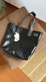 Bolso tipo tote bag negro con textura símil cuero de cocodrilo. Tiene doble asa corta, cierre superior y un dije colgante con forma de ala.