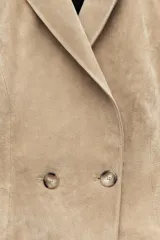 Blazer corto de cuero en color tostado, con cuello solapa, hombreras y manga larga. Presenta un diseño evasé en el bajo y un cierre frontal cruzado con dos botones.