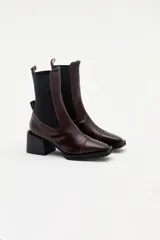 Bota tipo chelsea de cuero charolado color bordó, con caña media, elásticos laterales negros y taco cuadrado bajo.