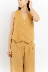 Chaleco de lino color mostaza con escote en V y cierre frontal de tres botones. Presenta costuras decorativas en contraste y un diseño de corte recto con terminación en punta.