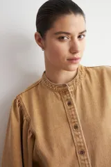 Camisa de jean color camel, de manga larga abullonada, con cuello mao y cierre frontal de botones metálicos.