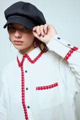Camisa blanca de algodón con detalles de puntilla roja en cuello, solapa frontal, bolsillo y puños. Presenta botones en color celeste.