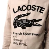 Camisa de manga larga Lacoste en color beige, con cuello abotonado y cierre frontal de botones. Presenta un estampado gráfico grande en el frente con el nombre de la marca, el logo del cocodrilo y la leyenda 'French Sportswear est. nineteen thirty three'. Incluye un pequeño logo bordado en el puño de la manga izquierda.