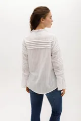 Camisa blanca de algodón con diseño romántico, cuello alto con volados, cierre frontal con botones nacarados y mangas largas con puños acampanados. Presenta detalles de encaje y franjas horizontales en toda la prenda.
