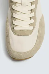 Zapatilla deportiva estilo barefoot, color marrón, con diseño de puntera ancha y redondeada. Confeccionada en una combinación de materiales textiles y detalles en serraje. Cuenta con suela fina de goma flexible y cordones en color crema.
