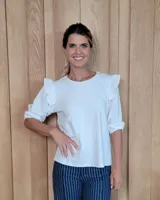 Remera blanca de cuello redondo con mangas tres cuartos que presentan volados en los hombros y puños elásticos.