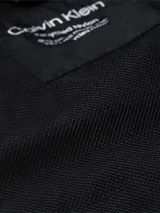 Campera negra con capucha de nailon reciclado, estilo blusón, con cierre de cremallera, bolsillos delanteros con cremallera, puños y dobladillo elásticos, tirador de cremallera con la marca e insignia con el logo de Calvin Klein en la manga.