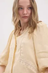 Camisa de lino color beige, de silueta romántica con escote a la base y mini volado. Cuenta con mangas 3/4 abullonadas con frunces y terminación en elástico. Posee detalles de puntillas negras en contraste en el frente y cierre frontal con botones ocultos bajo cartera.