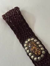 Cinto trenzado de cuero color chocolate con hebilla ovalada de metal envejecido, decorada con una piedra central jaspeada y tachas metálicas alrededor.