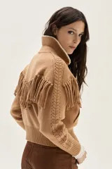 Bomber de tejido de punto color camel, con cuello de lana merino, cierre frontal, flecos decorativos en el frente y la espalda, y detalle de trenzas en las mangas.