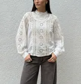 Camisa blanca de algodón con detalles de encaje y bordado calado en el frente y las mangas. Presenta cuello alto con volado, mangas largas abullonadas con puños y cierre de botones en la espalda.