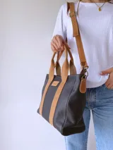Cartera de cuero negro con detalles en cuero color suela, incluyendo asas fijas y correa larga ajustable. Presenta una etiqueta frontal con logo grabado y cierre superior.