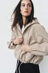 Campera estilo cazadora con capucha ajustable con cordones, cierre frontal de cremallera, mangas largas con puños elásticos, bolsillos delanteros de doble vivo y bajo ajustable con cordones. Presenta un diseño de cuadros en tonos beige y marrón.