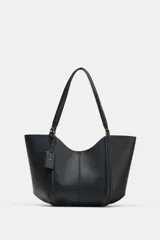 Bolso tipo shopper de hombro en color beige, con diseño estructurado, doble asa y detalle de pespuntes verticales en el cuerpo. Incluye un colgante decorativo tipo etiqueta y cierre imantado.