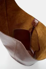 Bolso tipo saca de diseño asimétrico confeccionado en piel de color marrón. Cuenta con un asa de hombro ajustable y cierre mediante imán. Incluye una cartera con bolsillo extraíble en su interior.