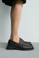 Mocasín de vestir color marrón oscuro, con empeine liso y detalle de antifaz. Presenta horma cuadrada y suela con ligero tacón geométrico a contraste.