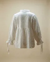 Camisa de algodón blanca con diseño holgado, cuello alto con volados, frente con tablas verticales y cierre frontal con botones. Presenta mangas largas amplias con puños ajustables mediante lazos.