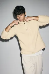 Campera deportiva de tejido sherpa color beige, con cuello elevado, cierre frontal completo, mangas raglán, bolsillo superior con cierre y bolsillos laterales. Presenta terminaciones elásticas en puños y ruedo.