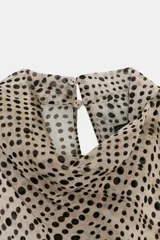 Blusa de cuello subido y manga sisa, confeccionada en tejido fluido semitransparente con estampado de lunares en tonos beige y negro. Presenta un diseño superpuesto tipo capa con caída asimétrica y cierre posterior con abertura y botón.