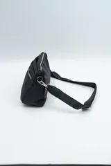 Bandolera de diseño compacto en color negro, confeccionada en material sintético resistente. Cuenta con un compartimento principal con cierre y un bolsillo frontal con cierre metálico visible. Incluye una correa larga ajustable y extraíble para llevar al hombro o cruzada.