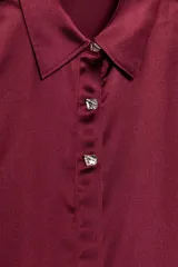 Camisa de cuello solapa y manga larga, confeccionada en tejido satinado con acabado brillante. Presenta cierre frontal con botones y bajo asimétrico.
