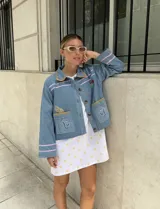 Chaqueta de jean celeste con bordados artesanales y a máquina. Presenta detalles de pespuntes ondulados en rosa y amarillo en el cuello, puños y bolsillos. Los bolsillos frontales tienen bordados de un limón y una figura marina estilizada.