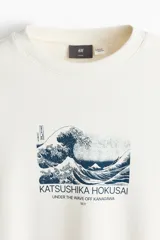 Buzo blanco off-white de corte holgado y hombros caídos, con cuello redondo, puños y bajo acanalados. Presenta un estampado frontal en el pecho inspirado en la obra de Katsushika Hokusai, 'La gran ola de Kanagawa'.