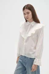 Camisa de algodón blanca con cuello camisero, cierre frontal de botones y volados bordados en el frente y hombros. Presenta mangas largas con puños acampanados y detalles bordados en el ruedo.