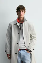 Trench coat de corte relajado, color gris claro, confeccionado en tejido técnico con mezcla de algodón. Presenta cuello solapa, manga larga con puño ajustable, bolsillos de doble vivo en cadera, bolsillo interior y abertura en el centro de la espalda. Cierre frontal con botonadura oculta.