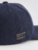 Gorra tipo baseball confeccionada en pana color azul marino, con parche rectangular lateral que incluye el logo de la marca Benson & Thomas.