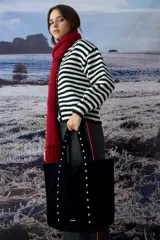 Cartera tipo shopper de gran tamaño confeccionada en material efecto gamuza color rojo. Presenta correas reforzadas decoradas con tachas metálicas plateadas y un dije colgante en forma de corazón con detalles de tachas. Cuenta con una placa metálica pequeña con logo en la parte inferior frontal.