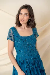 Vestido largo de tul color petróleo, con escote redondo y mangas cortas. Presenta un cuerpo bordado con lentejuelas al tono y una falda con movimiento decorada con apliques de hojas de tela.