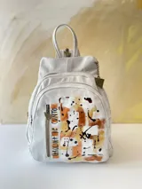 Mochila de cuero blanca con diseño artístico pintado a mano en tonos dorados, marrones y negros en el frente, múltiples bolsillos con cierre y herrajes de metal.
