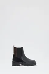 Bota tipo chelsea de caña media, con diseño combinado en cuero liso negro en la parte frontal y gamuza negra en el talón, con detalle de panel trasero en cuero marrón oscuro. Cuenta con elásticos laterales y suela track de goma negra.