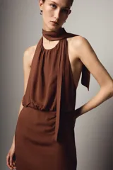 Vestido midi de color marrón con cuello halter y lazada en la nuca. Presenta un diseño con espalda descubierta, frunces en la cintura y una abertura lateral en la falda.