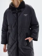 Tapado largo hasta las rodillas, color negro, con diseño acolchado de rombos. Presenta vivos a contratono y detalles en pana negra en el cuello y los bolsillos. Tiene cierre frontal con botones a presión, bolsillos delanteros y un cordón ajustable en la cintura. El forro es de algodón y lleva el logo "pastiche club" bordado en el pecho.