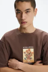 Remera marrón de algodón con cuello redondo, mangas cortas y hombros caídos, de corte holgado. Presenta un estampado frontal con la obra 'Los girasoles' de Van Gogh y el texto 'Sunflowers' en la parte superior y '-Van Gogh-' en la inferior.