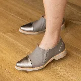 Zapato tipo mocasín de cuero vacuno en tono gris topo, con puntera reforzada en cuero metalizado color bronce. Presenta un diseño de corte lateral y suela plana con pequeña elevación en el talón.
