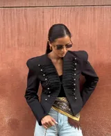 Chaqueta de estilo militar con estructura sastrera, hombros marcados y cuello mao. Presenta un diseño frontal con aplicaciones trenzadas y botones metálicos decorativos, de manga larga y corte corto.