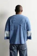 Camiseta boxy fit de color azul con efecto lavado vintage, cuello redondo y mangas por debajo del codo. Presenta un gran número '94' estampado en el frente y franjas horizontales en las mangas.