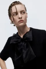 Saco de punto suave color negro, de manga corta y cuello redondo. Presenta cierre frontal con botones forrados y una moña decorativa de organza en la parte superior.
