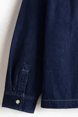 Sobrecamisa de denim de algodón rígido, color azul, con cuello inglés, bolsillos de parche en el pecho y en la parte inferior, y cierre frontal de botones metálicos.