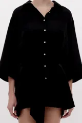 Camisa larga de satén negro, fluida, con cuello camisero, cierre frontal de botones dorados, mangas tres cuartos y lazo ajustable en la cintura.