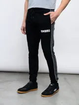 Pantalón deportivo Umbro modelo UNY, color negro, confeccionado en tejido de piqué. Presenta puños ajustados en los tobillos, cordón de ajuste en la cintura y una franja lateral gris oscuro ribeteada en blanco. El logo Umbro está estampado en blanco en el muslo izquierdo.