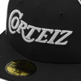 Gorra de béisbol de visera plana, colaboración entre Corteiz y New Era. Presenta un diseño bicolor en negro y gris con el logo de Corteiz bordado en el frente y el logo de New Era en el lateral.